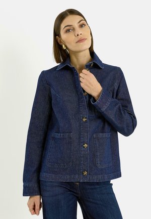 Mujer con cabello castaño liso, llevando aretes de aro dorados y una chaqueta vaquera azul oscuro con bolsillos frontales, abotonada hasta la mitad.