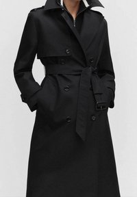 Czarny płaszcz trencz wykonany z gładkiej tkaniny, z podwójnym zapięciem na guziki, paskiem w talii, dużymi klapami i bocznymi kieszeniami.