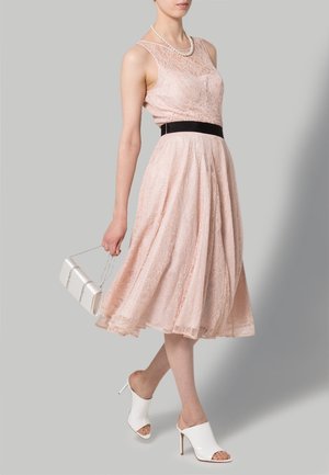 Femme portant une robe à manches courtes en dentelle rose avec une ceinture noire à la taille, un collier de perles, des mules blanches à talons hauts, et tenant une pochette blanche.