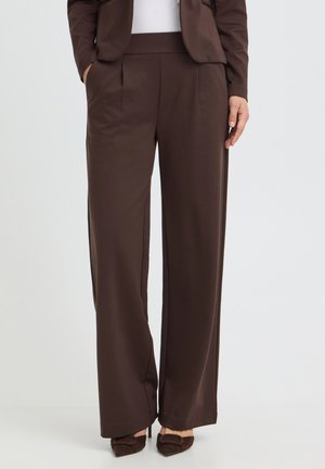 Personne portant un pantalon large de costume marron, un blazer assorti et des chaussures marron, debout les mains dans les poches devant un fond uni clair.
