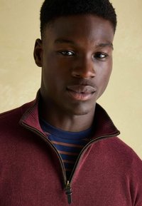 Bordový zipsový sveter s rebrovaným golierom, prekrytý námornícky pruhovanou košeľou. Má zlatý zips a hladkú textúru.