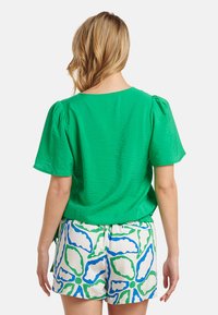 Grüner, strukturiert Oberteil mit kurzen Puffärmel, kombiniert mit leichten weißen Shorts, die ein blau-grünes Blumenmuster aufweisen.
