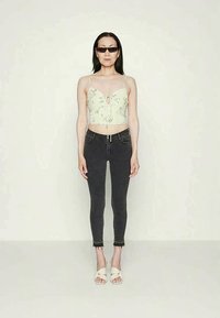 Mulher com cabelo longo e escuro, a usar óculos de sol, uma blusa floral curta, jeans negros justos com bainhas desfiadas e sandálias brancas de salto.
