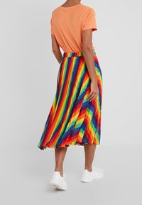 Jupe plissée colorée présentant des rayures verticales en rouge, orange, jaune, vert, bleu et violet ; associée à un haut simple orange et des baskets blanches.