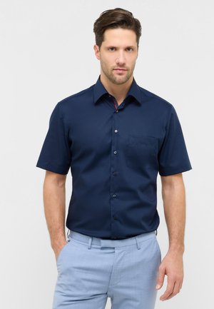 Mann mit kurzem braunem Haar, der ein marineblaues Kurzarmhemd mit Knöpfen und hellblaue Hosen trägt, steht mit einer Hand in der Tasche.