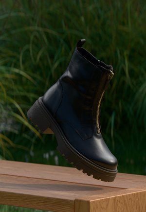Bottine en cuir noir avec fermeture éclair dorée à l’avant et semelle épaisse crantée flottant au-dessus d’une surface en bois en extérieur.