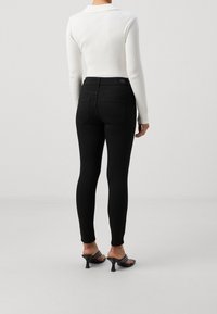 Jeans negros ajustados de cintura alta, con una textura suave y dos bolsillos traseros, combinados con una camiseta blanca de manga larga con canalé y tacones negros.