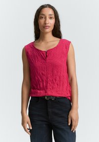 Haut sans manches rouge froissé avec un détail de bouton au niveau du col. Associé à un jean en denim foncé et une ceinture noire.