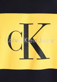La camiseta de Calvin Klein Jeans presenta un diseño en bloques de color amarillo y negro con un gran logo negro "CK" y texto de la marca en blanco. Fabricada en algodón.