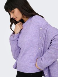Kvinna med mörkt hår som bär en mjuk lavendelfärgad tröja och matchande kofta med knappar, justerar kragen med ena handen mot en ljus bakgrund.