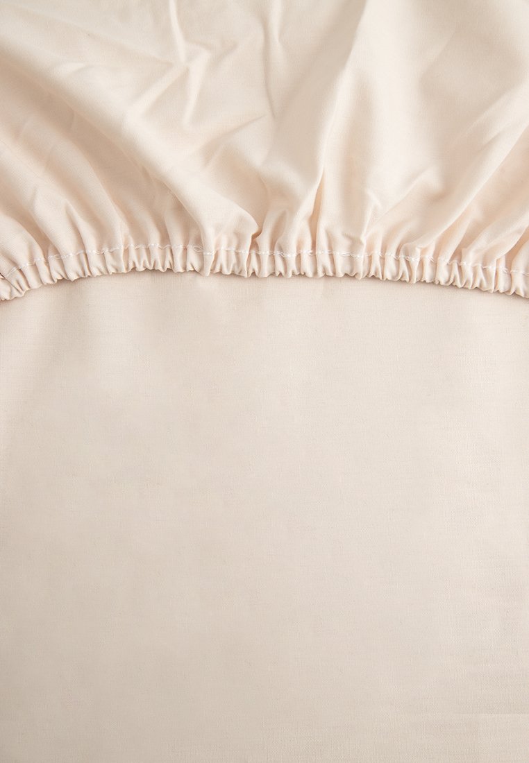 Housse de matelas beige ajustée avec un bord élastique, étendue sur un coin de matelas, montrant un tissu lisse et une couture élastique froncée.