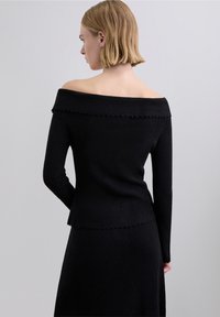 Czarny sweter z odsłoniętymi ramionami, z długimi rękawami, teksturowaną tkaniną i falowanym wykończeniem u dołu, widziany od tyłu.