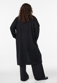 Lange schwarze Tunika mit langen Ärmeln, aus weichem Stoff; kombiniert mit schwarzen, weit geschnittenen Hosen und flachen schwarzen Schuhen. Rückansicht gezeigt.