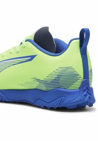 Zapatilla deportiva verde lima con acentos en azul, superficie texturizada y un patrón de piso geométrico; cuenta con una pestaña en el talón para facilitar su uso.