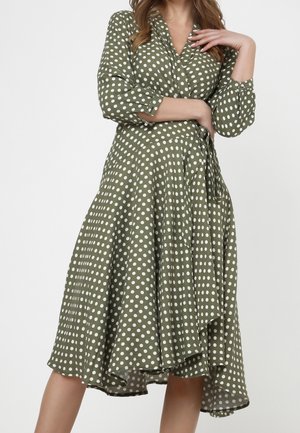 Žena v olivově zelených zavinovacích šatech s bílými puntíky, se tříčtvrtečními rukávy a asymetrickým lemem, pózující s jednou rukou blízko obličeje.