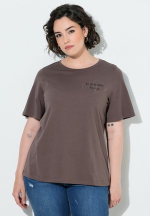 Frau mit lockigem dunklem Haar, die ein braunes T-Shirt mit dem Text "Irresistibly Yours" und blaue Jeans trägt, steht vor einem weißen Hintergrund.