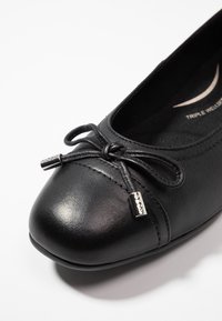 Svart lädersko med platt sula och rund tå, dekorativ rosett och metalldetalj. Har en strukturerad yta och stoppad innersula.