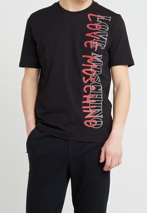 Homme portant un t-shirt noir à manches courtes avec le texte vertical "LOVE MOSCHINO" en rouge et blanc sur le côté droit, associé à un pantalon noir.