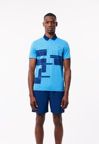Blå polo skjorta med en kontrasterande marinblå krage, prydd med geometriska mönster i marinblått. Bärs med matchande marinblå shorts.