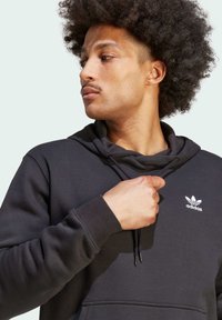 Felpa con cappuccio nera realizzata in tessuto morbido. Presenta una tasca a canguro frontale, polsini a costine e un logo Adidas ricamato in bianco.