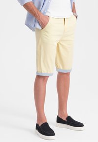 Pantaloni corti di cotone gialli con risvolti, dotati di tasche laterali. Abbinati a scarpe nere slip-on e a una camicia azzurro chiaro con bottoni.