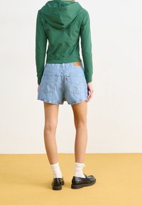 Grön kort hoodie, ljusblå jeansshorts, svarta skor, vita strumpor. Noterbara detaljer inkluderar fickor och en läderpatch på shortsen.