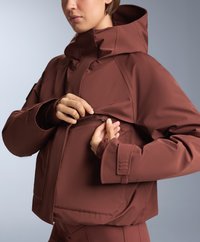 Rjava kapuca z zadrgo na strani in elastičnimi manšeti. Tkanina se zdi gladka s strukturirano prileganjem in minimalnimi oblikovalskimi podrobnostmi.