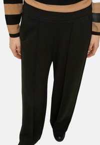 Pantaloni neri a gamba larga con una texture liscia, caratterizzati da una vita piatta e dettagli sulle cuciture laterali, abbinati a una maglia a righe.