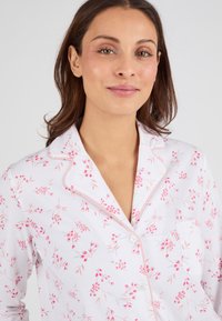 Haut de pyjama en tissu blanc avec un imprimé floral rose, doté d'un col à encolure décolletée, d'une poche avant et d'une fermeture à boutons avec des détails en passepoil rose.