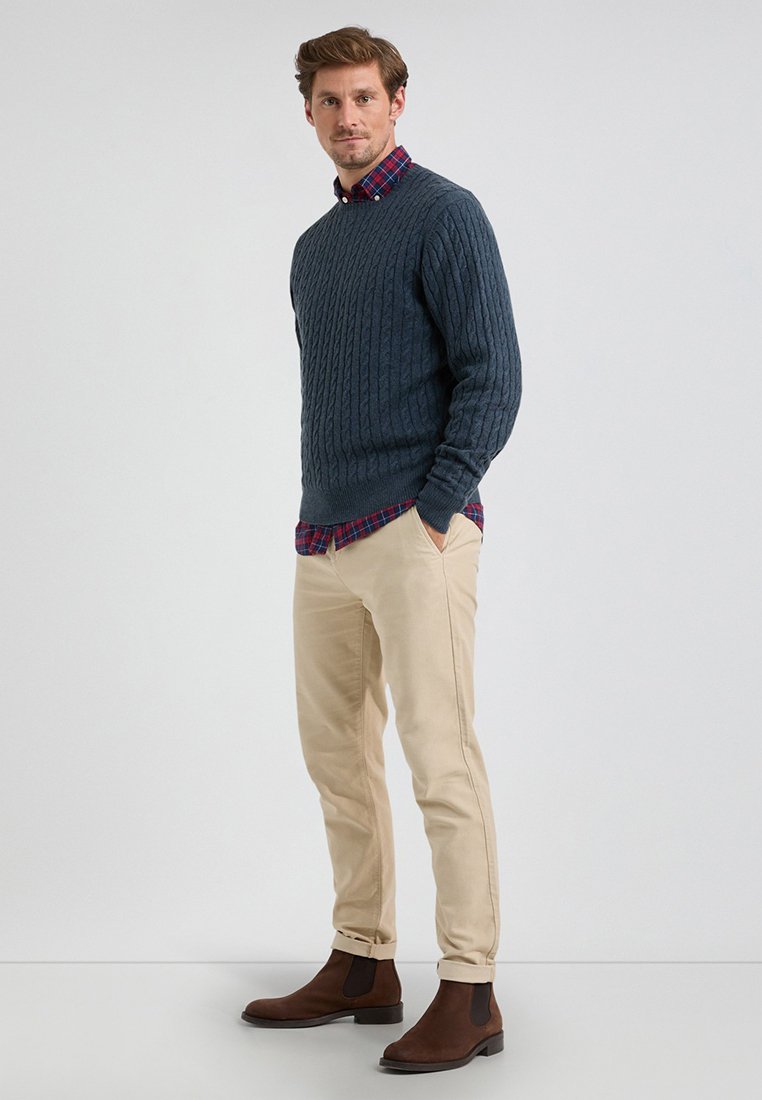 Donkerblauwe kabeltrui gedragen over een rood-blauw geruit shirt, gecombineerd met beige chinos en bruine Chelsea boots.
