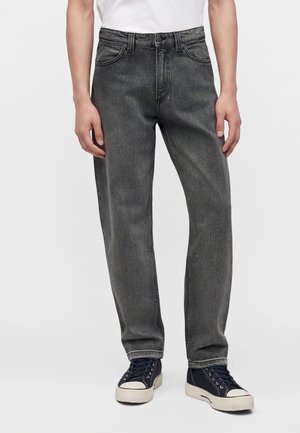 Personne portant un jean noir délavé, coupe droite, et des baskets noires basses avec semelles et lacets blancs, debout devant un fond uni.