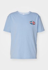 Camiseta de algodón azul claro con mangas cortas. Presenta un logo bordado circular con el texto "TOMMY" y detalles en rojo, blanco y azul marino.