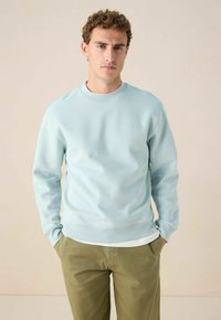Sweat-shirt en coton bleu clair avec col rond, poignets à côtes et ourlet. Présente un petit logo sur la poitrine. Associé à un pantalon vert olive.