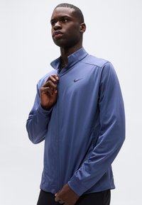 Άνδρας που φοράει μπλε αθλητικό μπουφάν με φερμουάρ Nike, τραβώντας το φερμουάρ προς τα πάνω, με φόντο ένα απλό ανοιχτό γκρι φόντο.