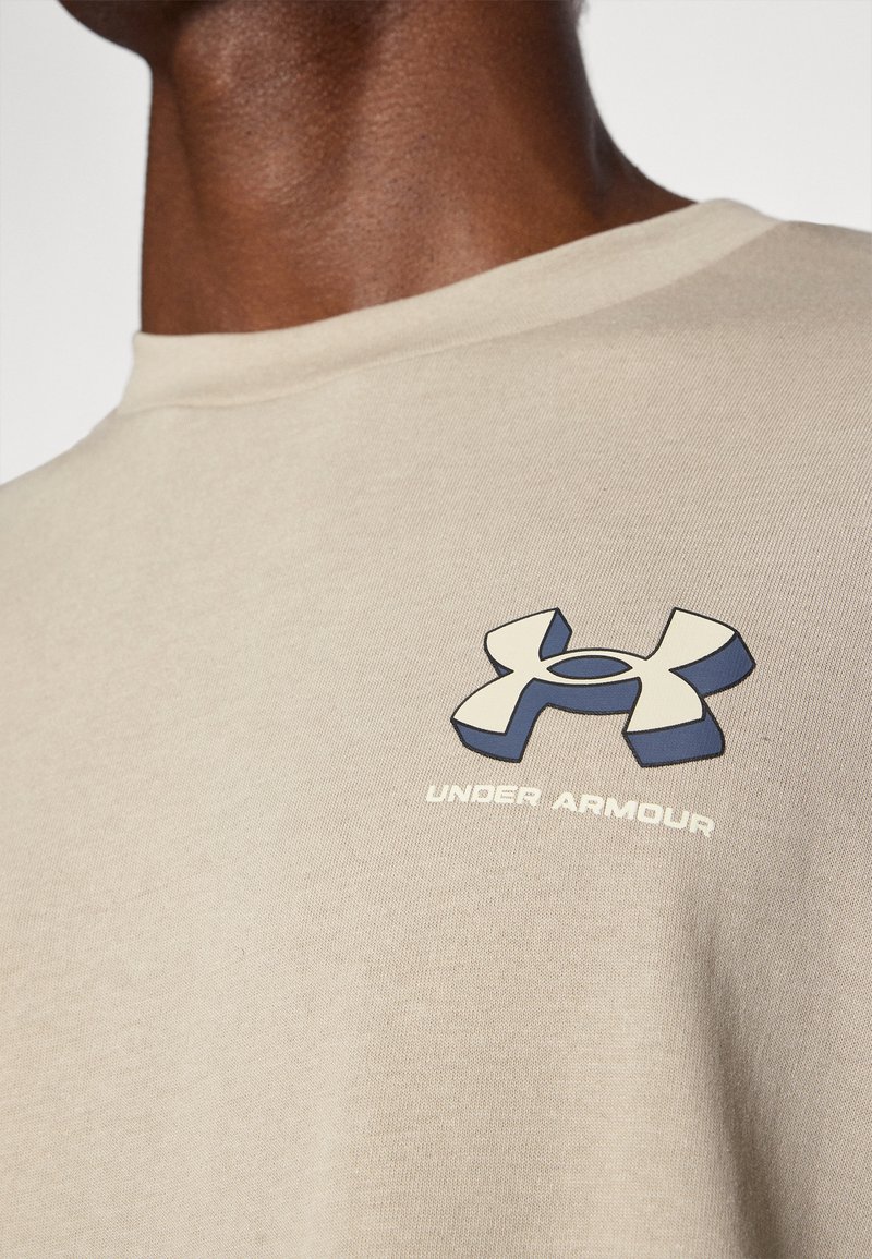 T-shirt in cotone beige con collo rotondo. Presenta un logo Under Armour blu scuro sul petto e il testo "UNDER ARMOUR" sotto il logo.