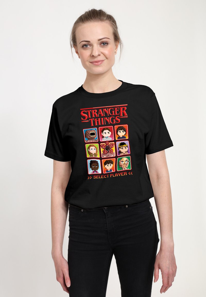 Černé tričko s krátkým rukávem s pixel art designem postav ze seriálu "Stranger Things" a textem "SELECT PLAYER" níže.