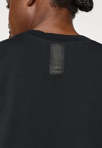 Schwarzes T-Shirt mit glatter Textur und einem auffälligen gewebten Logolabel am hinteren Halsausschnitt. Einfaches Design, keine zusätzlichen Muster oder Akzente.