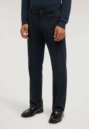 Uomo che indossa scarpe eleganti nere, pantaloni blu navy e una camicia a maniche lunghe scura e strutturata, in piedi davanti a uno sfondo semplice.
