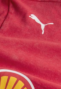 Czerwona koszulka z tkaniny z białym logo Puma oraz okrągłą, pomarańczowo-żółtą grafiką z wzorami promieni słonecznych. Miękka tekstura, casualowy styl.