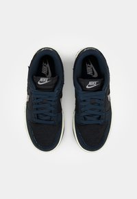 Pár tmavě modrých Nike tenisek s tkaničkami, bílými mezipodešvami a viditelnými logy Nike na jazyku a stélkách, fotografováno shora na bílém pozadí.