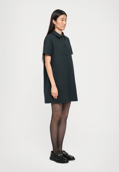Tummanvihreä lyhythihainen mekko, jossa on napitettu etuosa, kaulus ja sivutaskut. Mallilla on päällään mustat loaferit ja tummat sukkahousut.