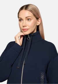 Navy zip-up sweatshirt with high collar, drawstrings, and metallic accents on pocket. Soft fabric, relaxed fit, and long sleeves.