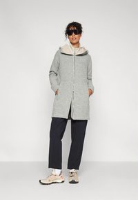 Siv plašč s kapuco z zadrgo spredaj, z lahkim bež podlogo. Kombiniran z mornarsko modrimi hlačami in bež supergami. Gladek tekstura, sproščen kroj.