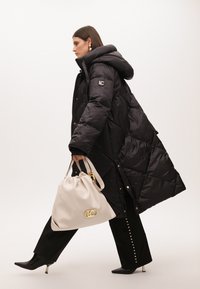 Černý prošívaný dlouhý kabát s kapucí a logem, doplněný bílou taškou a černými vysokými botami na podpatku s metalickými detaily.