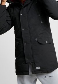 Zwarte geïsoleerde parka met drukknoopsluiting, twee voorzakken en een verstelbare capuchon; gecombineerd met grijze sweatpants met een logo.