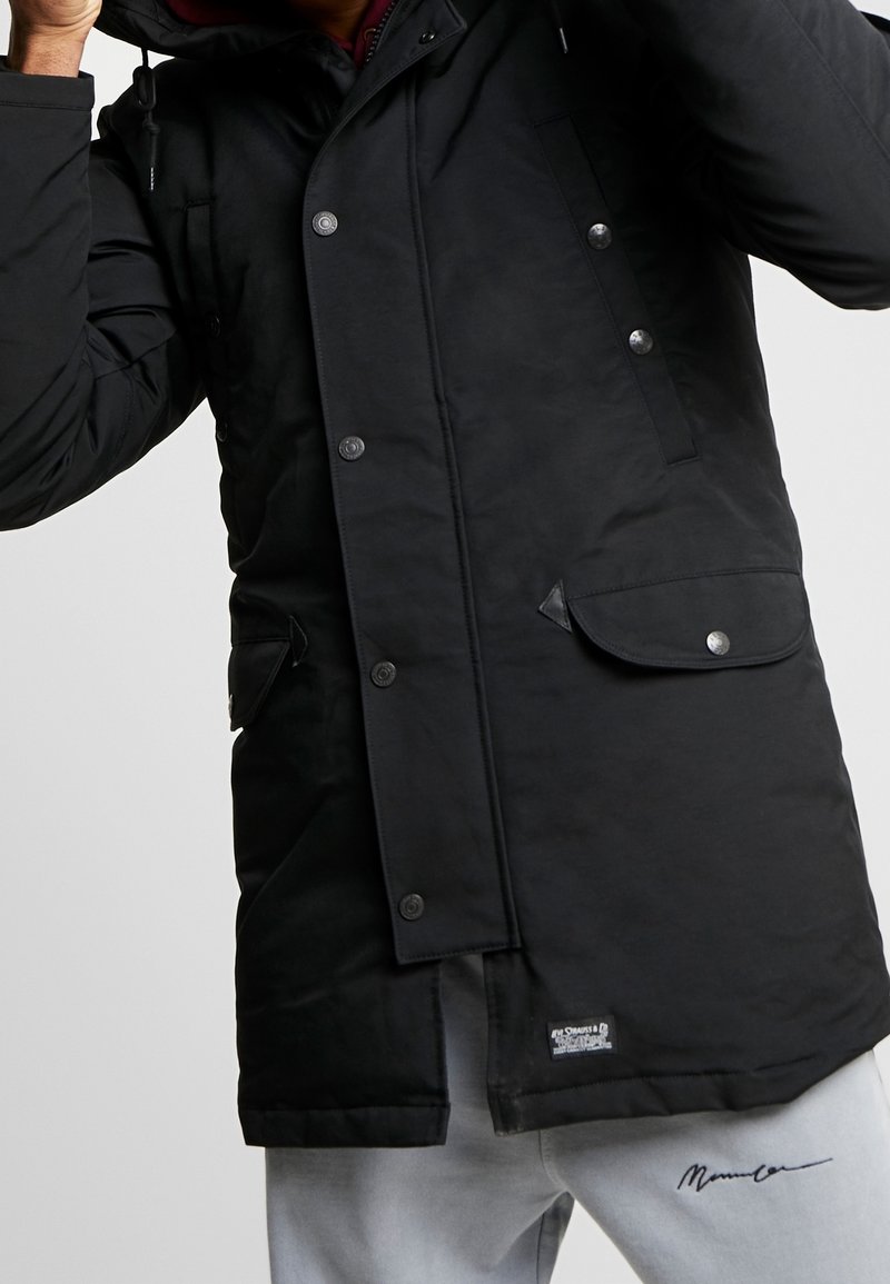 Zwarte geïsoleerde parka met drukknoopsluiting, twee voorzakken en een verstelbare capuchon; gecombineerd met grijze sweatpants met een logo.
