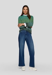 Frau mit verschränkten Armen, die einen grün-blauen Strickpullover, ein weißes Hemd, blaue Jeans und weiße Turnschuhe trägt, vor einem einfarbigen Hintergrund.