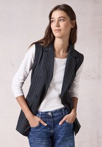 Navy-blau gestreifte ärmellose Weste mit Reverskragen, zwei vorderen Taschen, kombiniert mit einem weißen Langarmhemd und blauen Jeans.