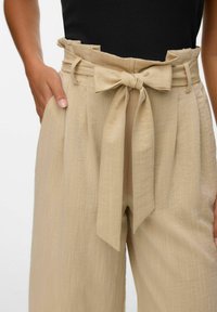 Beige högmidjade byxor med texturerad yta, med vida ben och en knuten rosett vid midjan. Fickor på sidorna.
