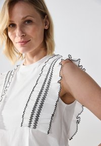 White top featuring ruffled sleeves with black embroidery. The design includes multiple horizontal lines and contrasting textures.
